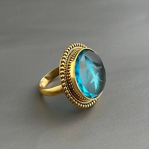 May include: A gold ring featuring a large blue gemstone set in a bezel setting. The gemstone has a carved design of a bird.