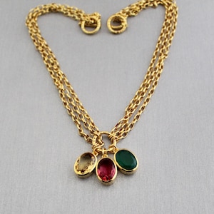 Puede incluir: Un collar de cadena de múltiples hilos en tono dorado con tres dijes ovalados. Los dijes incluyen una piedra amarilla translúcida, una piedra roja oscura y una piedra verde sólida, engastadas en marcos de metal dorado en un anillo central.