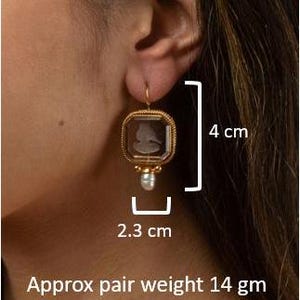 May include: Gold tone earrings with a square glass cameo featuring a profile portrait and a pearl accent. The earrings measure 4 inches in length and 2.3 inches in width. The pair weighs approximately 14 grams.
