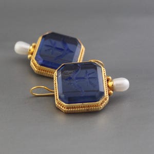 May include: A pair of gold earrings with blue glass insets. Each earring features a square-shaped blue glass stone with a gold frame and a small pearl hanging from the bottom.