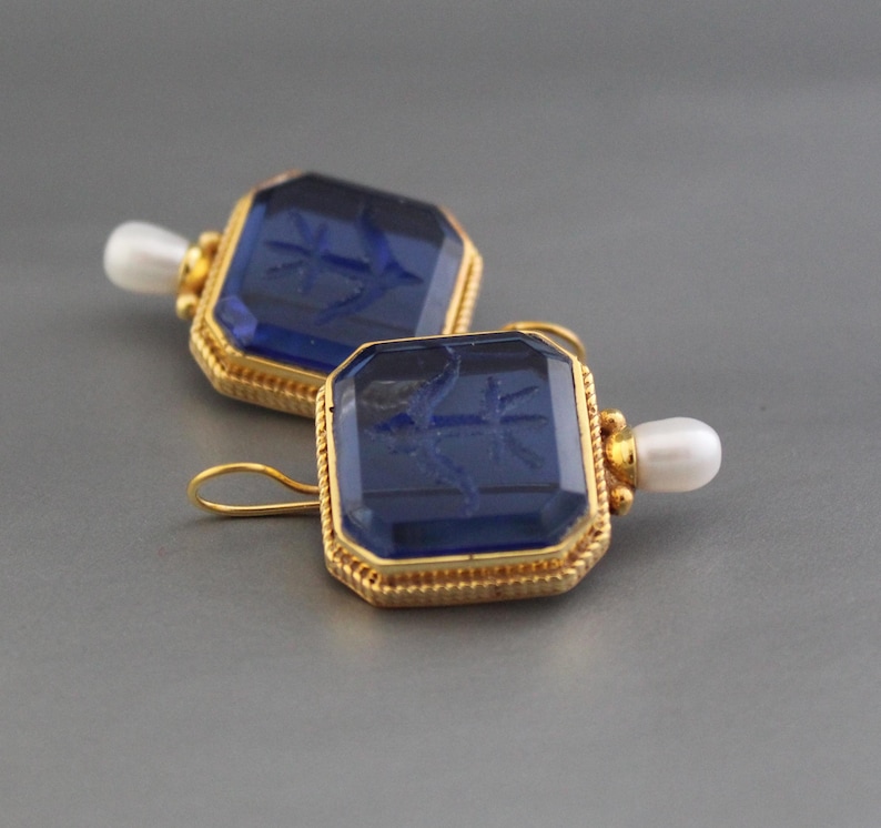 May include: A pair of gold earrings with blue glass insets. Each earring features a square-shaped blue glass stone with a gold frame and a small pearl hanging from the bottom.