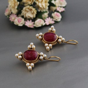 Peut inclure: Une paire de boucles d'oreilles en or avec des accents de pierres précieuses rouges et des embellissements de perles blanches. Les boucles d'oreilles ont un design suspendu avec une petite boucle en bas.