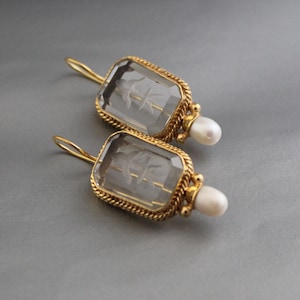 Puede incluir: Un par de pendientes dorados con colgantes rectangulares de cristal transparente. Cada colgante presenta un diseño grabado de un pájaro. Los pendientes tienen un borde en forma de cuerda y una gota de perla blanca. El fondo es gris liso.