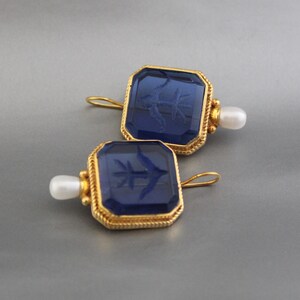 May include: A pair of gold earrings with blue glass insets and a pearl accent. The glass insets are square and have a carved design.