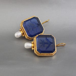 May include: A pair of gold earrings with blue glass insets. The glass is etched with a floral design and the earrings are accented with small white pearls.