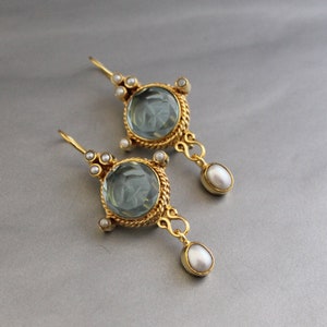 K&ouml;nnte beinhalten: Ein Paar goldene Ohrringe mit einem runden, hellblauen Edelstein und einem Perlenanh&auml;nger. Die Ohrringe haben ein detailliertes, verziertes Design.