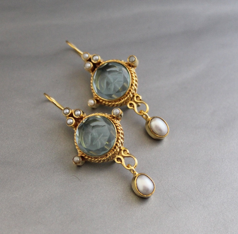 K&ouml;nnte beinhalten: Ein Paar goldene Ohrringe mit einem runden, hellblauen Edelstein und einem Perlenanh&auml;nger. Die Ohrringe haben ein detailliertes, verziertes Design.