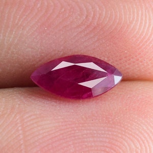 Può includere: Gemma di rubino taglio marquise, di colore rosso intenso. La gemma sfaccettata ha una superficie lucida con sfaccettature bianche riflettenti. Il rubino è tenuto tra le dita, mostrando il suo colore e la sua forma.
