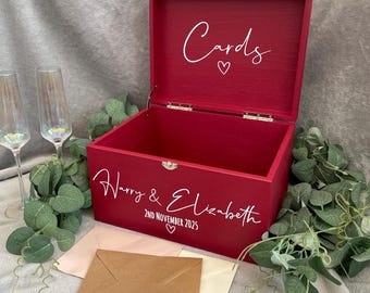 May include: A red wooden box with a hinged lid. The lid is open and says "Cards" with a heart symbol. The box is decorated with the text "Harry & Elizabeth" and "2nd November 2025" with a heart symbol. There are three envelopes on the table in front of the box.