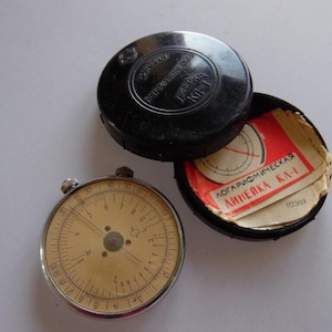 Può includere: Un regolo calcolatore vintage con un quadrante circolare e una cornice in metallo, accanto a una custodia nera aperta, che rivela una pila di carte rosse e bianche. La custodia è etichettata in russo.