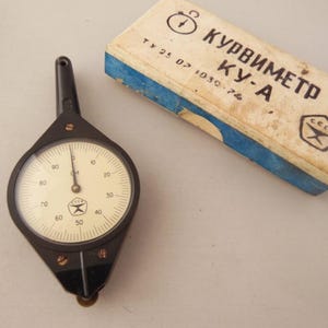 May include: A vintage black and white mechanical curve meter with a circular dial, marked in increments from 0 to 90. The device is next to a rectangular box with Cyrillic text and blue accents.