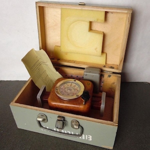 Puede incluir: Una caja de madera abierta, pintada de verde claro por fuera y con el interior de madera natural. En el interior, un instrumento de madera marrón con un indicador circular y un botón negro descansa sobre una base roja. También hay un papel y una caja gris.