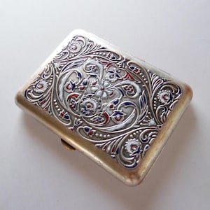 Könnte beinhalten: Eine silberne Zigarettenetui mit einem verzierten Blumendesign aus roter, weißer und blauer Emaille. Das rechteckige Etui hat abgerundete Kanten und einen Schnappverschluss. Das detaillierte Muster bedeckt die gesamte Oberseite.