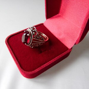 May include: A silver ring with a large, oval-shaped red gemstone set in a filigree design. The ring is displayed in a red velvet ring box.