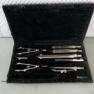 May include: An open black case containing a set of silver drafting tools. The set includes several compasses and other drawing instruments, with red and black accents. The case has a velvet lining.