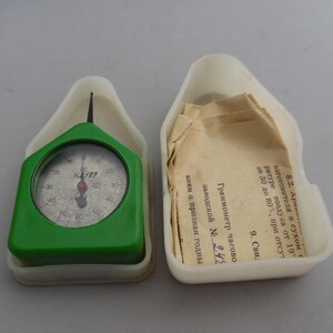 Könnte beinhalten: Ein grünes und weißes mechanisches Messgerät im Vintage-Stil mit einem kreisförmigen Zifferblatt, das Zahlen und eine Nadel anzeigt. Das Messgerät befindet sich in einem weißen Kunststoffgehäuse, neben einem offenen Gehäuse mit Papieren.