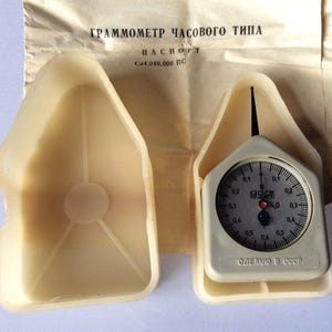 Könnte beinhalten: Ein cremefarbenes Vintage-Gramm-Messgerät mit rundem Zifferblatt und schwarzem Zeiger, untergebracht in einem passenden Kunststoffgehäuse. Das Zifferblatt zeigt Zahlen und Markierungen. Der Text "MADE IN USSR" ist sichtbar.
