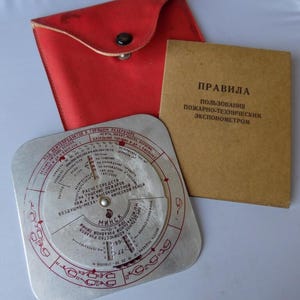 Może przedstawiać: Zabytkowe urządzenie przeciwpożarowe z okrągłą tarczą, czerwonymi oznaczeniami i broszurą. Urządzeniu towarzyszy czerwone skórzane etui z zatrzaskiem. Broszura zawiera tekst w języku obcym.