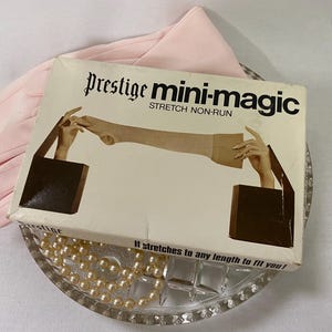 May include: A vintage box of "Prestige mini-magic" stretch non-run hosiery, with an illustration of a leg being stretched. The box is on a glass dish with a pearl necklace and a pair of light pink gloves.