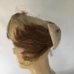 May include: A vintage hat with a cream-colored satin bow and a delicate net veil. The hat features a combination of cream and brown feathers, creating a textured and elegant design. This classic headwear is perfect for special occasions.