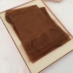 May include: A pair of brown nylon stockings folded inside a pink-trimmed box. The stockings have a patterned design near the top and are a solid, neutral color. The box is open, revealing the contents.
