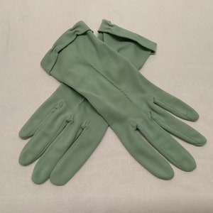 May include: A pair of sage green gloves. The gloves are made of a smooth fabric and have a classic design. The gloves have a seam running down the back of each finger and a folded cuff at the wrist. The gloves are laid out on a light-coloured surface.