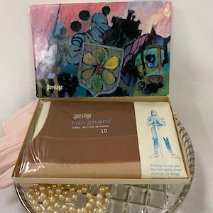 May include: Vintage "Prestige Run Guard" knee-action nylon stockings in a box. The box features a colorful illustration of knights and a shield. The stockings are a light brown color. The box is displayed on a glass dish with pearls.