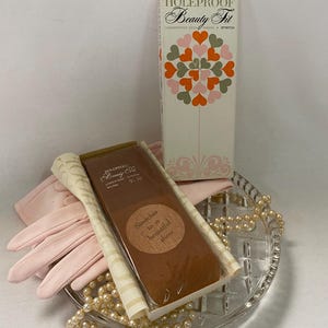 May include: A vintage display featuring a box of "HOLEPROOF Beauty Fit" hosiery, a pair of pink gloves, and a strand of faux pearls. The hosiery box has a heart-shaped design. The items are arranged on a clear, patterned glass dish.