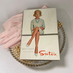 May include: A vintage box of "Sutex" seamless mesh hosiery, featuring a seated woman in a light blue dress and white heels. The box is displayed with pink gloves and a glass dish with pearls.