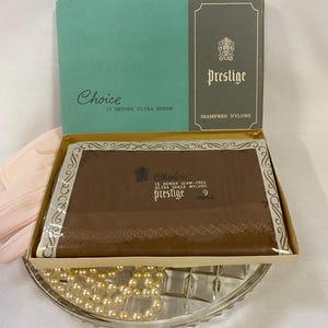 May include: A vintage box of "Choice" 15 denier ultra sheer "Prestige" seamfree nylon stockings. The box is brown and teal with gold trim. The stockings are a dark brown color. The box is sitting on a glass dish with pearls.