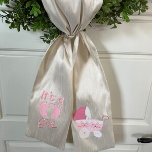 May include: A white satin ribbon with pink embroidery. The ribbon says "It's a Girl" with a pair of pink footprints and a pink baby carriage.