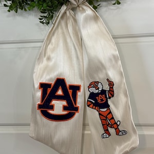 May include: A white satin ribbon with an embroidered Auburn University logo and mascot. The logo is a navy blue "AU" with an orange outline. The mascot is an orange and black tiger wearing a navy blue shirt with the "AU" logo.