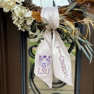 May include: White fabric door wreath bow with purple and gold LSU Tigers logo and text.