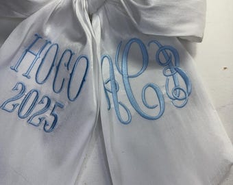 May include: A large white satin bow with light blue embroidered text. The text includes "HOCO 2025" and a monogram. The bow is tied in the center, with long, flowing tails. The embroidery is detailed and elegant.