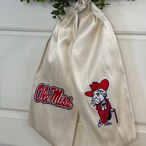 May include: A white satin bow tie with an embroidered Ole Miss logo and a cartoon mascot wearing a red hat and holding a cane.
