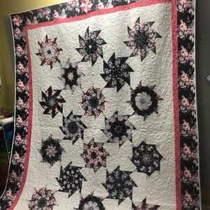 May include: A white quilt with a black and pink floral pattern. The quilt is made up of a series of star-shaped blocks, each with a different floral design. The quilt is edged with a pink and white border.