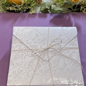 May include: A stack of white envelopes with a floral pattern, tied with twine. The envelopes are on a purple surface, with a floral arrangement in the background.
