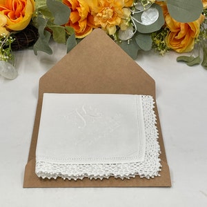 May include: White handkerchief with embroidered initials and delicate lace trim, presented in a brown envelope. The handkerchief features a monogram and text. The background includes yellow and green floral arrangement.