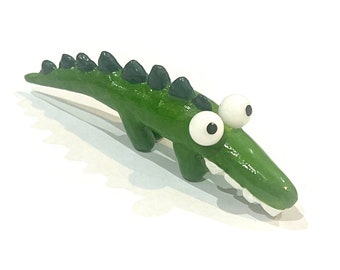 May include: A green alligator figurine with black spines and large white eyes. The alligator has a toothy grin and is made of clay.