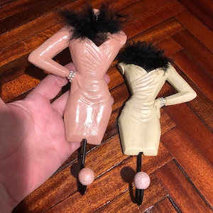 May include: Two decorative hooks shaped like women in dresses. The hooks are painted in a light pink and cream color. Each hook has a black feather boa around the neck and a pearl bracelet on one wrist.