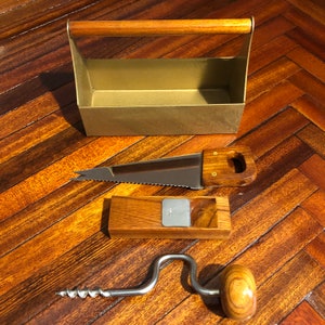 May include: A set of four vintage woodworking tools, including a small saw, a wooden plane, a hand drill, and a metal toolbox with a wooden handle. The tools are arranged on a wooden surface.