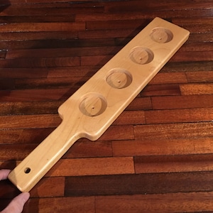 May include: A light-colored wooden paddle-shaped serving tray with four circular indentations. The tray has a handle with a hole at the end. The wood grain is visible, and the tray appears to be new.