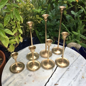 May include: Set of eight gold metal candle holders with varying heights and designs. The candle holders are arranged on a white marble surface.