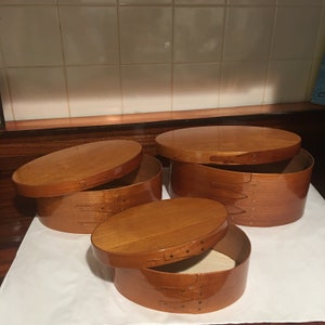 May include: Three wooden oval boxes with lids. The boxes are made of light brown wood and have a glossy finish. The lids are slightly ajar, revealing the interior of the boxes.