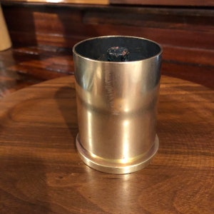 May include: A gold-colored metal cup with a black top. The cup is sitting on a wooden surface.