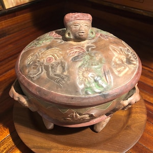 May include: A terracotta pot with a lid, decorated with colorful figures and designs. The pot has three legs and two handles. The lid features a human face with a headdress.