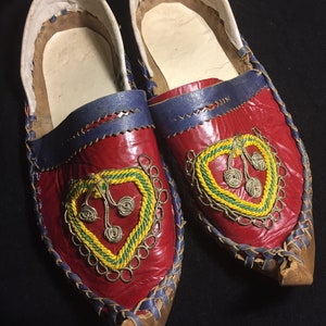 May include: Pair of red leather slippers with blue trim and intricate gold, green, and silver embroidery. The slippers have a pointed toe and a turned-up heel.