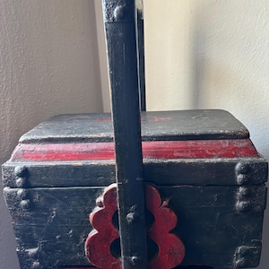 May include: A rectangular wooden box with a black finish and red accents, featuring a tall, fixed vertical handle. The box has a hinged lid, visible metal rivets along the edges, and decorative red scalloped brackets at the base of the handle.