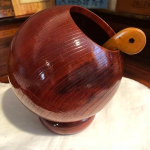 May include: A wooden bowl with a dark red stain and a matching wooden spoon. The bowl is round and has a small base. The spoon is curved and has a hole in the handle.