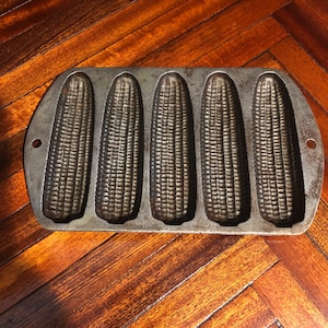 May include: A vintage cast iron cornbread pan with five individual molds. The pan is made of dark gray metal and has a slightly textured surface.